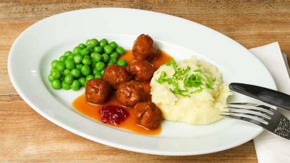 Köttbullar angerichtet auf einem Teller mit Erbsen, Kartoffelpüree, Rahmsoße und Preiselbeeren.