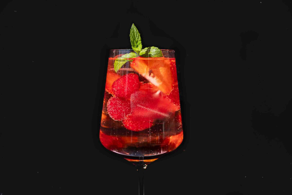 Lillet Wild Berry im Glas mit Eis, Erdbeeren, Himbeeren und Minze vor schwarzem Hintergrund