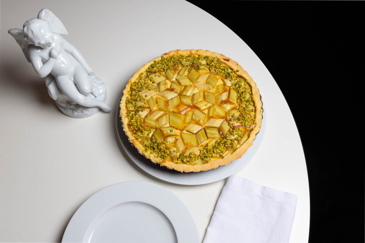 Rhabarber-Tarte mit Pistazienrand auf weißem Tisch neben Porzellan-Amor