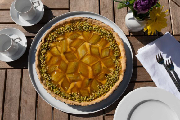 Rhabarbertarte mit Pistazien auf einem Holztisch im Garten, gedeckt mit Espressotassen und Blumenvase